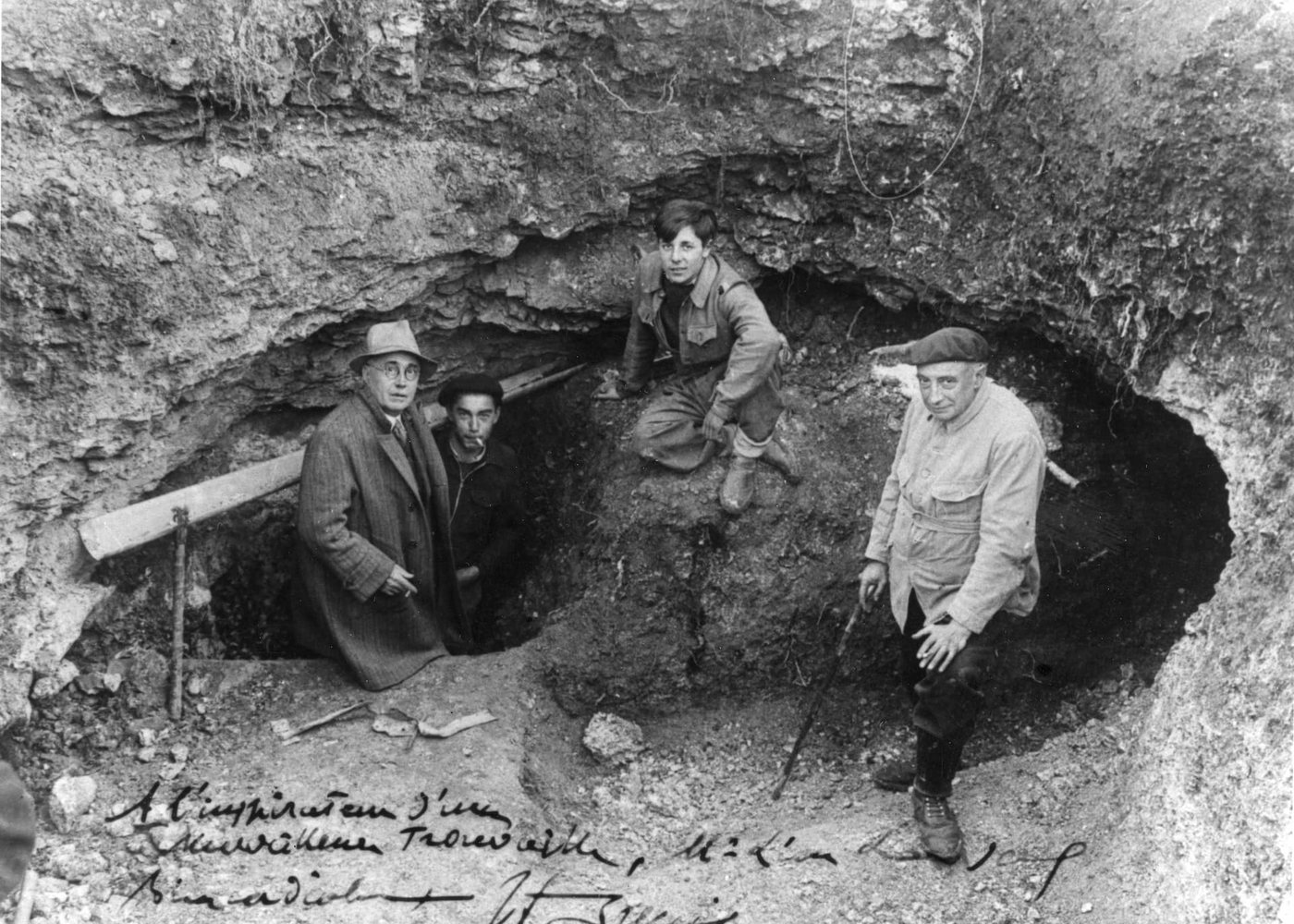 The entrance to Lascaux cave in September 1940: three young men and a man in a coat standing around the hole opened in the slope, on a dedicated print