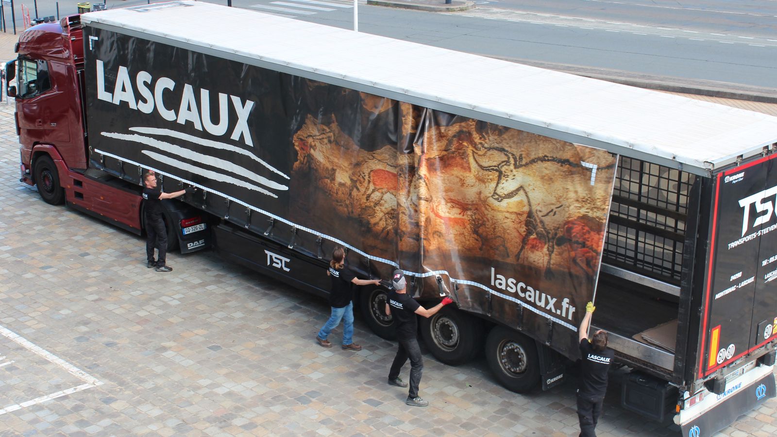The exhibition’s truck being unloaded on a square, its tarpaulin printed with the Hall of the Bulls